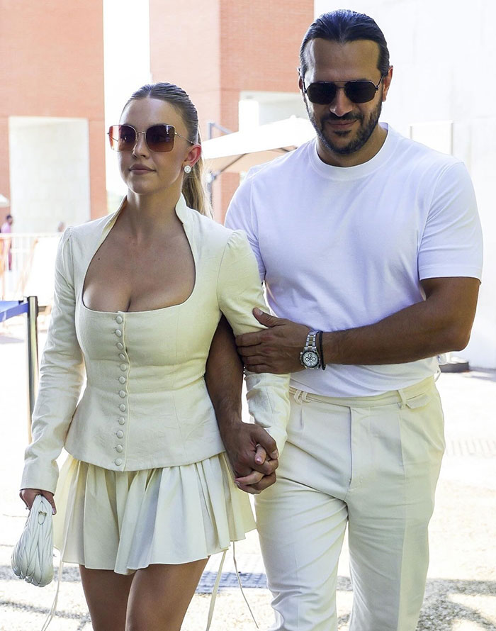 Sydney Sweeney is dressed in a cream-colored, button-up jacket with a matching pleated skirt, holding a small white bag. She wears sunglasses and has her hair tied back. Jonathan Davino walks beside her in a white T-shirt and cream pants, also wearing sunglasses. They hold hands as they walk together outdoors on a sunny day. Sydney Sweeney is dressed in a cream-colored, button-up jacket with a matching pleated skirt, holding a small white bag. She wears sunglasses and has her hair tied back. Jonathan Davino walks beside her in a white T-shirt and cream pants, also wearing sunglasses. They hold hands as they walk together outdoors on a sunny day.