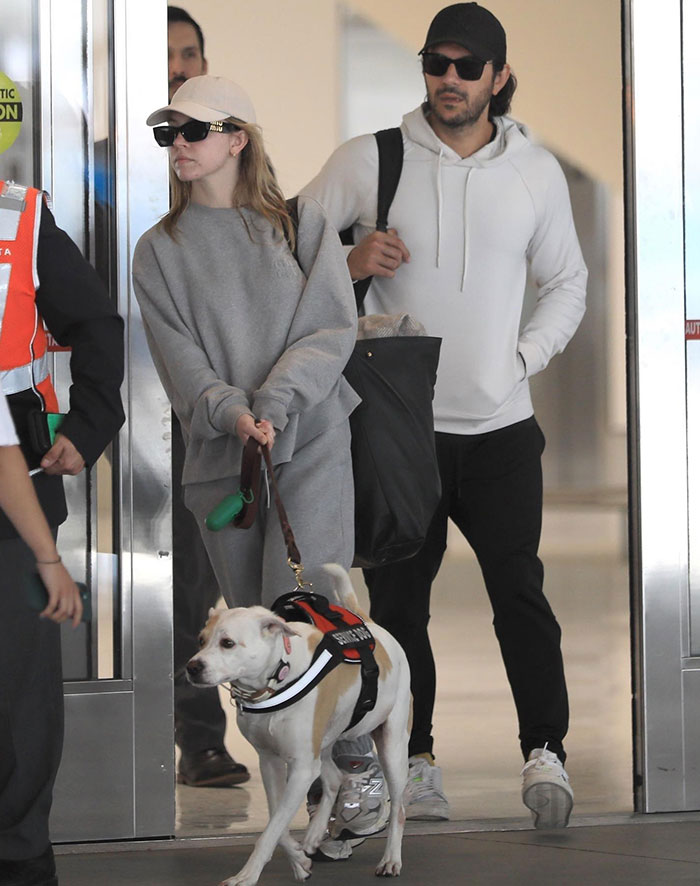 Sydney Sweeney, dressed in a gray sweatshirt and pants with a beige baseball cap and sunglasses, holds the leash of a white dog wearing a harness. Beside her, Jonathan Davino is dressed in a white hoodie, black pants, and a black cap, carrying a large bag over his shoulder. They appear to be at an airport or public indoor space, walking together with their dog. Sydney Sweeney, dressed in a gray sweatshirt and pants with a beige baseball cap and sunglasses, holds the leash of a white dog wearing a harness. Beside her, Jonathan Davino is dressed in a white hoodie, black pants, and a black cap, carrying a large bag over his shoulder. They appear to be at an airport or public indoor space, walking together with their dog.