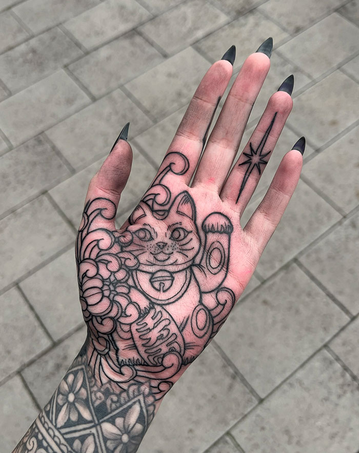 Hand tattoo featuring a black ink design of a lucky cat and patterns.