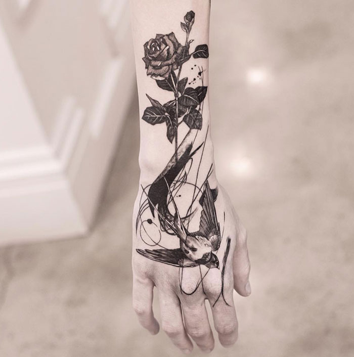 Black ink tattoo of a rose and bird on a woman's arm.