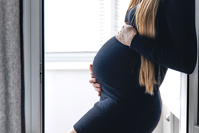 A young pregnant woman in a black dress stands near a window, touching her belly.