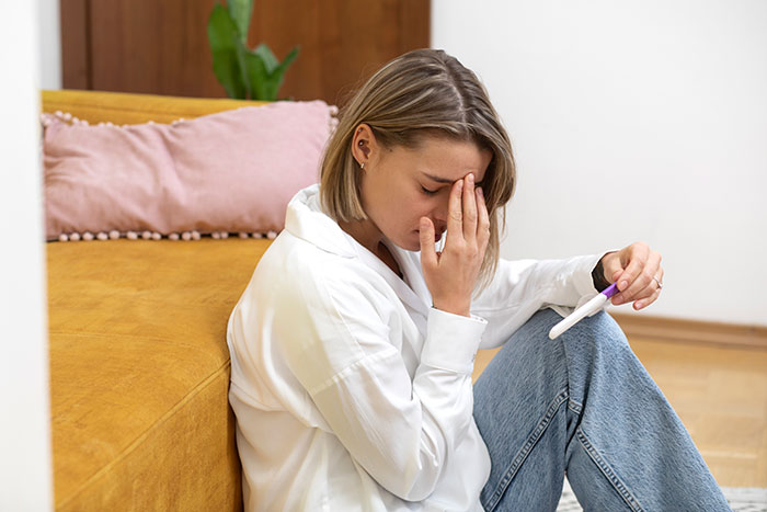 Teen girl sitting on the floor, distressed, holding a pregnancy test in a living room setting.