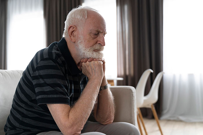 Elderly man seated, deep in thought, in a brightly lit room.