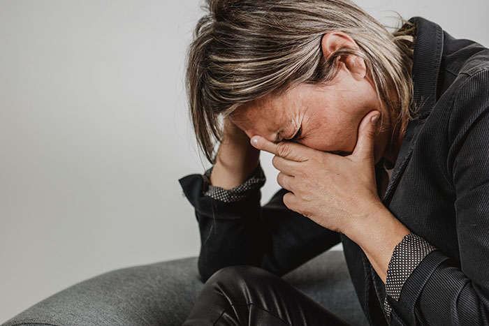 A woman sitting on a gray sofa, showing a painful expression, with her hand covering her face.