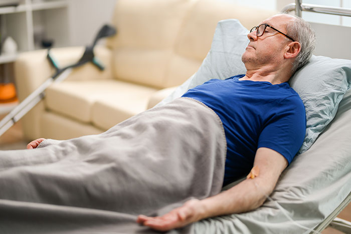 Elderly man resting in a hospital bed, wearing a blue shirt, with crutches in the background.