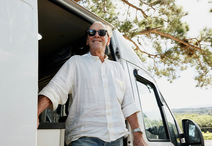 Man in sunglasses smiling, standing at the door of a van in a scenic area.