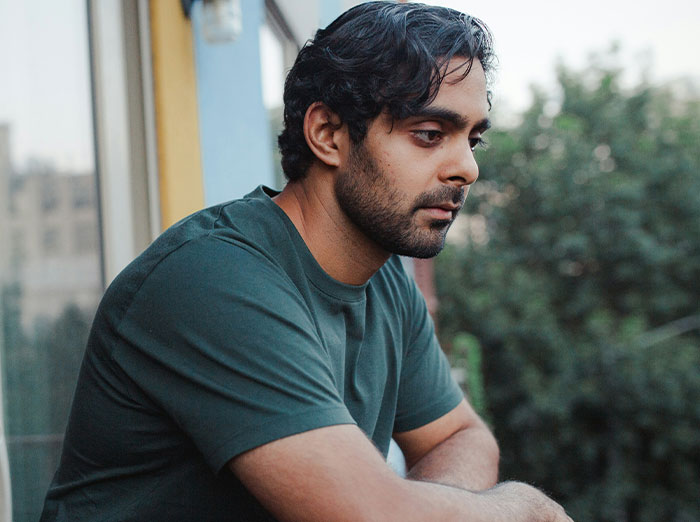 Man in a green shirt looks thoughtful, leaning on a balcony, with trees in the background.
