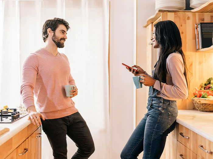 Man and woman chatting in a kitchen, both holding mugs, amidst a bright and cozy setting.