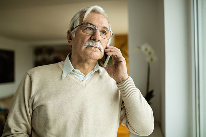 Elderly man wearing glasses, standing by a window, talking on a phone, looking thoughtful about family will. Elderly man wearing glasses, standing by a window, talking on a phone, looking thoughtful about family will.