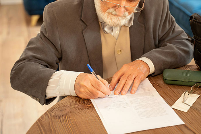 An elderly man in a suit signing a will at a wooden table, focusing intently on the document. An elderly man in a suit signing a will at a wooden table, focusing intently on the document.