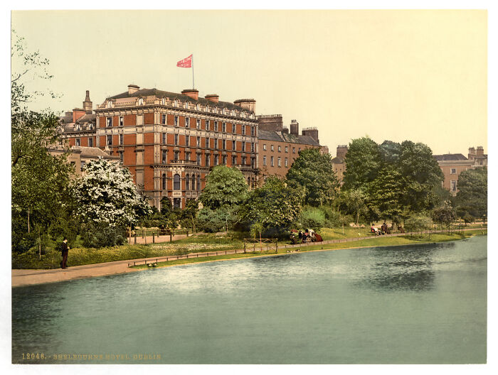Oldest color photo of 19th-century building by a river, surrounded by trees and people on a sunny day.