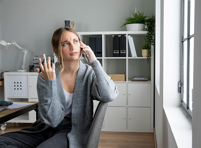 Woman looking frustrated while on the phone, holding a gesture of disbelief in a home office setting.