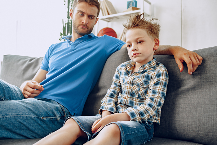 Man in blue polo looks concerned while sitting beside a boy on a sofa, reflecting tension in a family relationship.