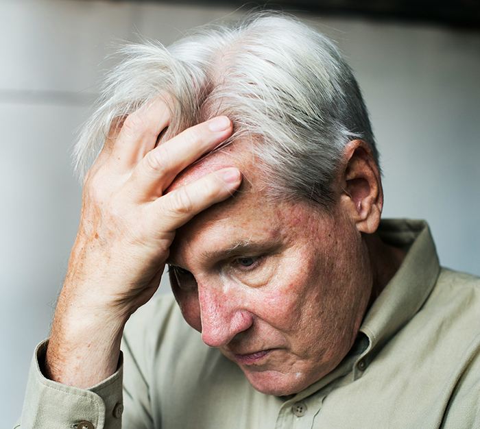 Older man looking upset, touching his head, wearing a beige shirt.
