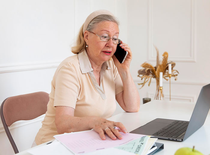 An elderly woman on a phone call, seated at a desk with a laptop and papers, discussing a company decision.