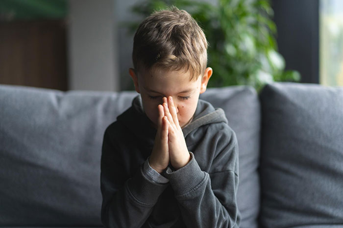 Young boy with clasped hands sitting on a gray sofa, highlighting family village dynamics.