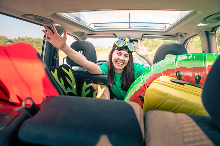 Woman happily packing car with vacation gear, waving from backseat.