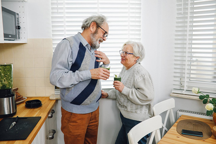 Elderly couple in a kitchen, smiling and holding drinks, representing in-laws visiting unexpectedly on vacation.