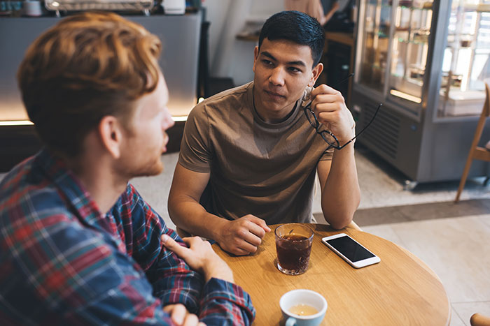 Two men at a caf&eacute; table, one holding glasses and listening, illustrating communication and cultural judgment.
