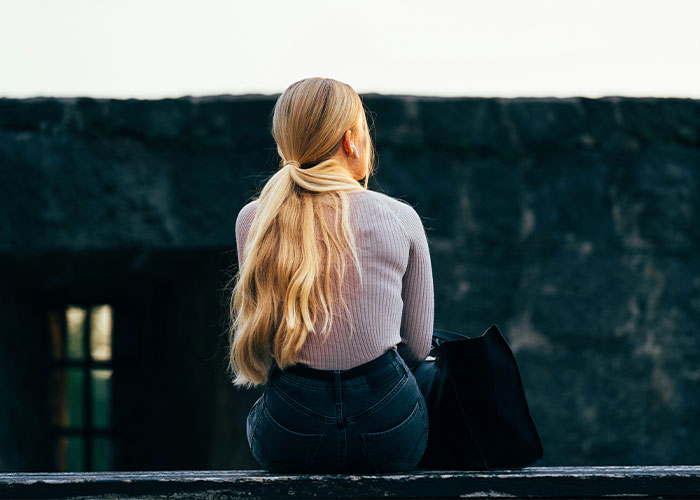 Person with long blond hair sitting outdoors, facing away, contemplating spilled secrets.