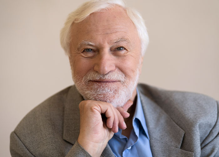 Elderly man in a gray suit smiling, hand on chin, conveying a sense of wisdom and reflection on secrets shared.