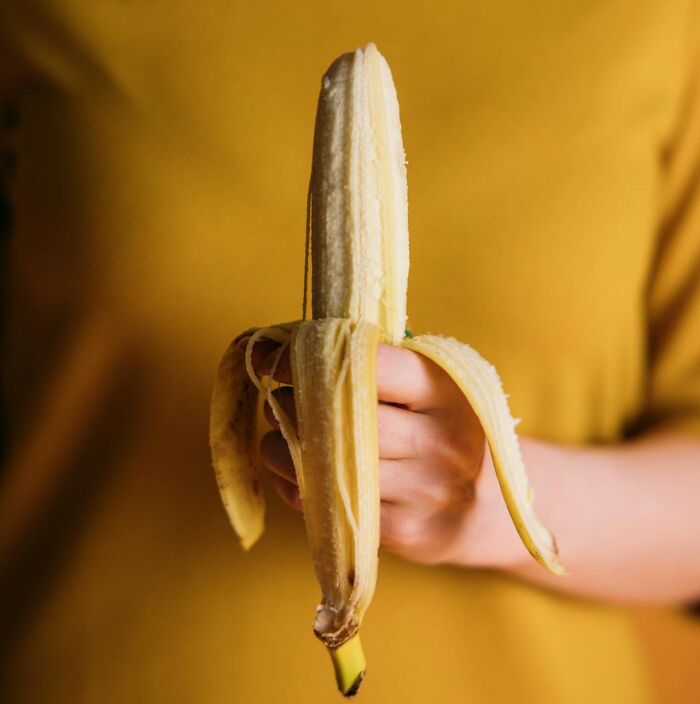 Person holding a partially peeled banana, standing against a yellow background.