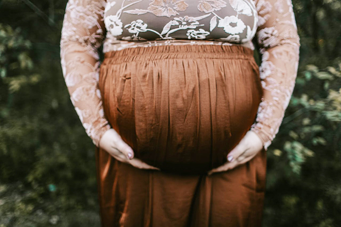Pregnant woman holding her belly, wearing a floral top and brown skirt, surrounded by greenery. Pregnant woman holding her belly, wearing a floral top and brown skirt, surrounded by greenery.