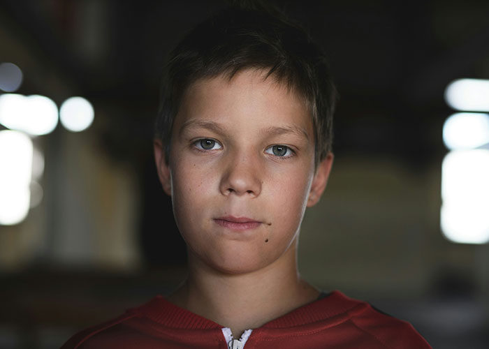 A young boy in a dimly lit room, looking directly at the camera with a neutral expression.