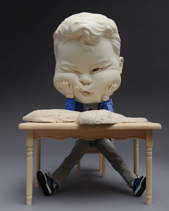 Realistic and surreal sculptural art depicting a child with an exaggerated head and expression, sitting at a table.