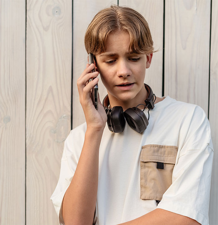 Teenager with headphones around neck, talking on phone, against wooden background.