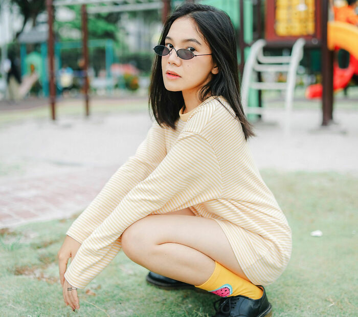 A woman in sunglasses crouches outdoors in a striped sweater dress, highlighting controversial fashion trends.