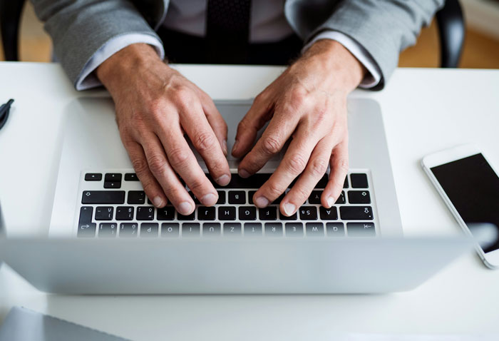Hands typing on a laptop keyboard representing reasons people have been fired, with a smartphone beside it on a desk.