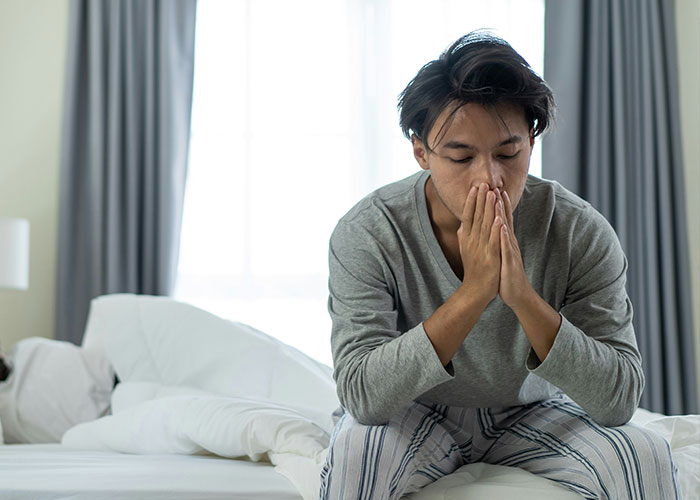 A man sitting on a bed looking regretful, hands clasped in front of his face.