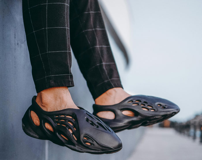 Feet wearing trendy black shoes with cut-out design, paired with checkered pants, against a city backdrop.