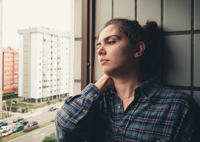 Person in plaid shirt, looking out a window with a reflective expression; urban buildings in the background.
