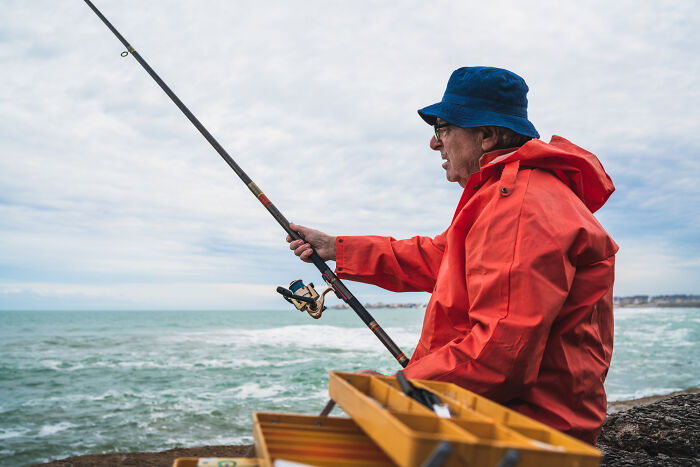 A man in a red raincoat and blue hat fishing by the sea, reflecting peaceful trends for 2025.