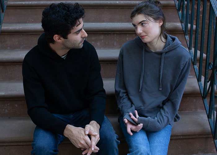 Two people sitting on steps, sharing secretive conversation, wearing casual hoodies and jeans.