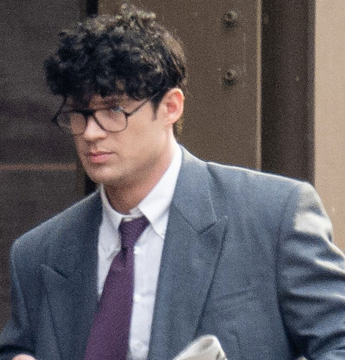 Man with curly hair and glasses in a gray suit and purple tie, possibly discussing fashion trends.