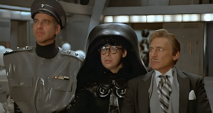 Three characters in a movie scene with comedic elements, wearing distinct costumes: an officer, a figure in dark helmet, and a man in a suit.