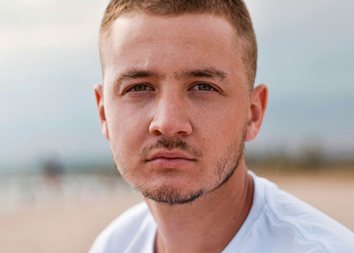 A man with short hair and a white shirt looking directly at the camera on a beach.