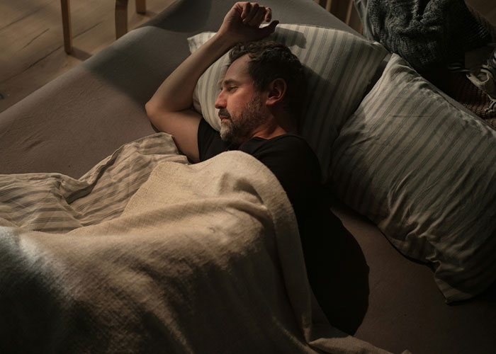 A man sleeping on a couch under a blanket, appearing thoughtful or regretful after revealing secrets.