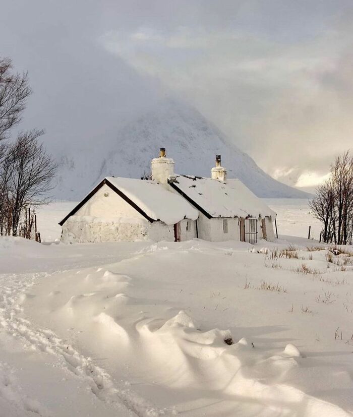 Snow-covered Scottish cottage in a serene winter landscape with distant mountains and bare trees.