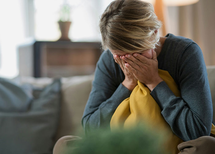 Person regretting a secret, sitting on a couch, covering their face with hands, embracing a yellow pillow.