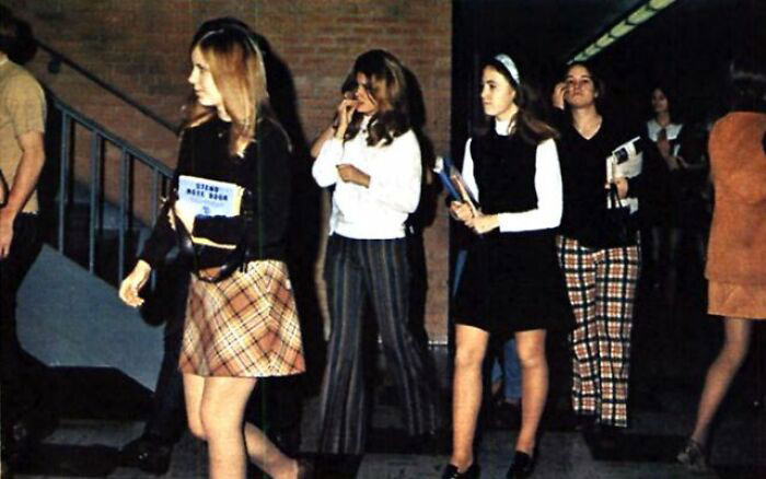 Students in 1970s high school fashion walk through a hallway, featuring plaid skirts and striped pants.