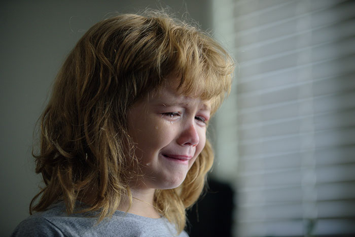 Child crying near window blinds, expressing sadness, related to affair child and Christmas gift context. Child crying near window blinds, expressing sadness, related to affair child and Christmas gift context.