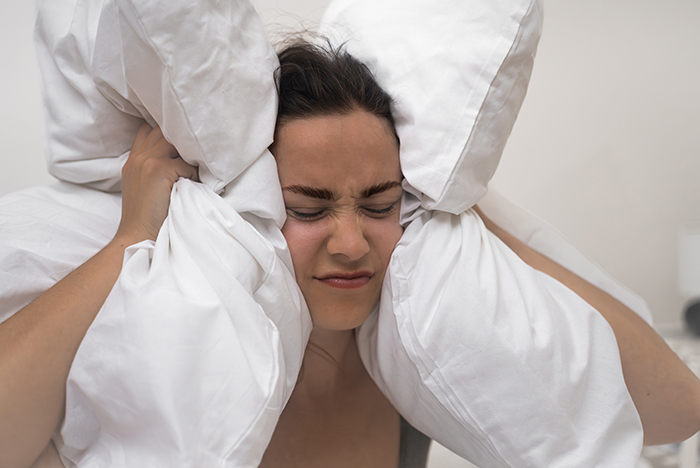 Woman covering ears with pillows, blocking noise, expressing frustration.
