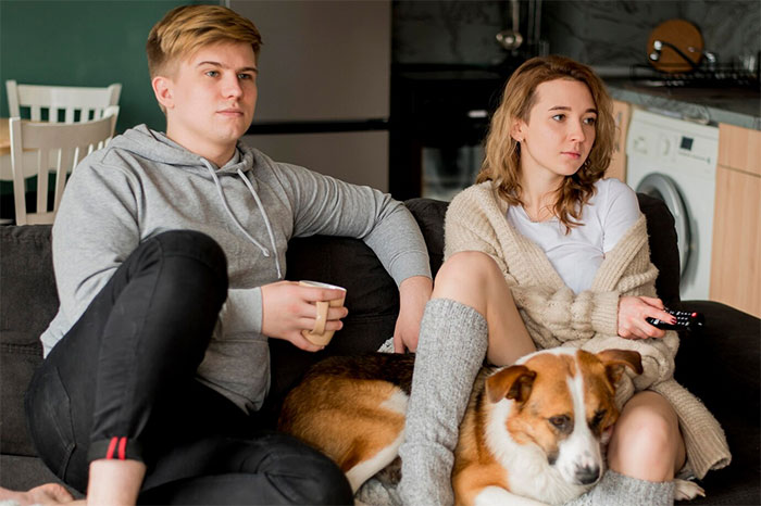 Couple sits on couch with dog, woman holds remote; themes of babysitting, infidelity, and vindictiveness are implied.