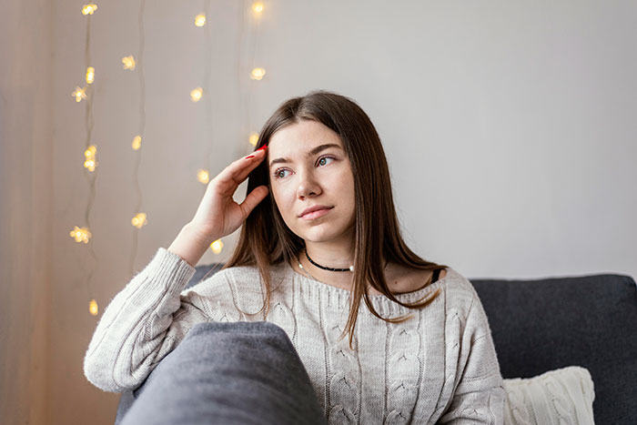 Woman sitting on a gray couch, reflecting, wearing a cream sweater with fairy lights in the background.