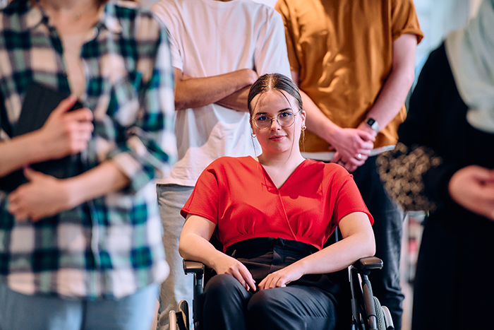 Woman in a red shirt sitting in a wheelchair, surrounded by people standing, depicting misunderstanding and disbelief.