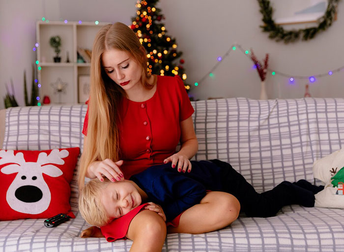 Mom in red dress, sitting on a couch, gently stroking a child's hair, Christmas decor in the background.